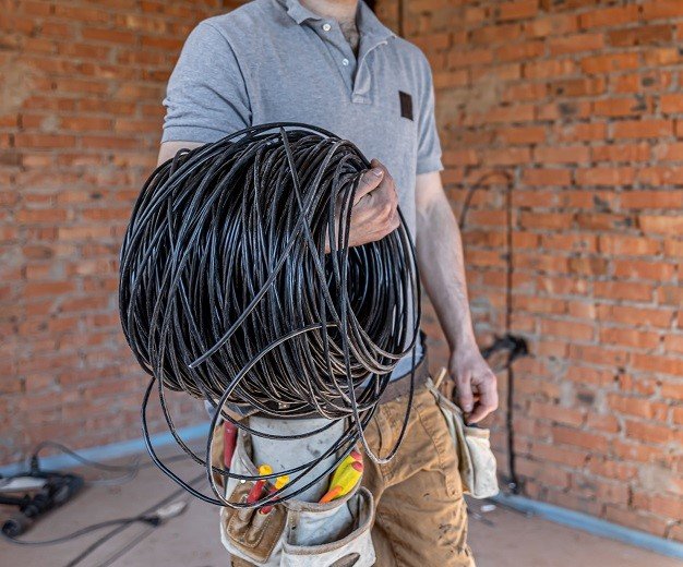 An electrician in a hard hat looks at the wall while holding an electric cable.